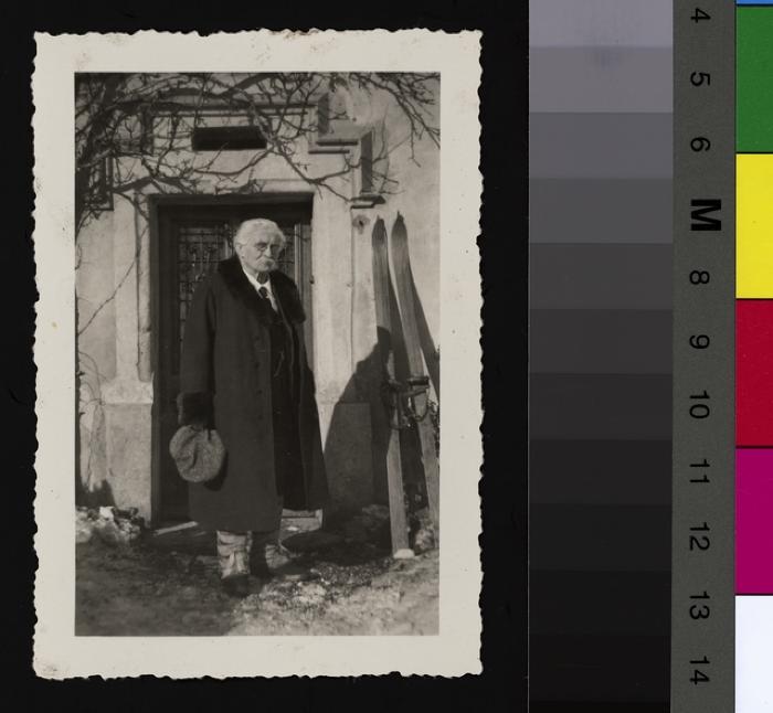 Portrait du Prof. Dr. César Roux âgé. Il pose devant la porte d'une maison. A sa gauche, des skis.